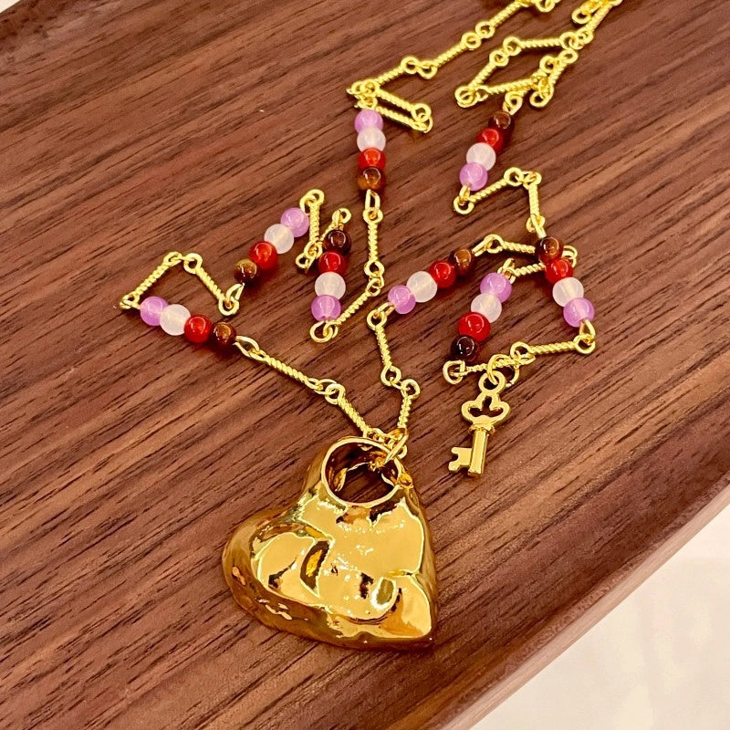 Collana lunga con pendente a cuore traforato color oro e texture particolare, con girocollo di perline di cristallo e pietra naturale.